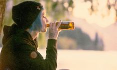 a woman drinking from a beer bottle while sitting on a bench in front of a tree