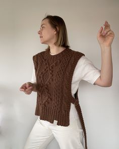 a woman in white pants and a brown knitted sweater is holding her hands up