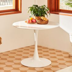 a white table with fruit and bread on it in front of two windows next to a potted plant