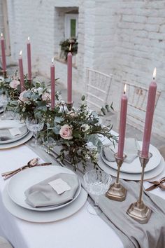 the table is set with white plates and silverware, pink candles, and greenery