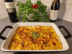 a casserole dish on a table with bottles of wine and a potted plant