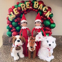 three elfs and two dogs are standing in front of a christmas wreath with the words we re back on it