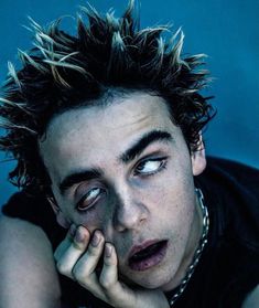 a young man with spiked hair and piercings looks at the camera while holding his hand to his face