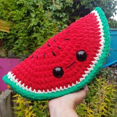 a hand holding up a crocheted watermelon cushion in front of some bushes