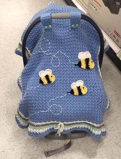 a blue crocheted baby seat cover with two bees on it, sitting next to a cardboard box