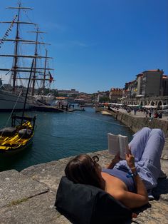 a woman reading a book while sitting on the edge of a pier next to a boat