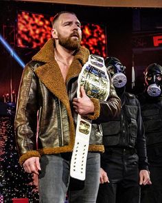 a man in a leather jacket holding a white wrestling belt with two other men standing behind him