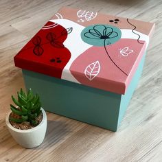 a small potted plant sitting next to a painted box on the floor in front of a wooden table