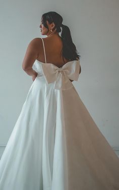 a woman in a white dress with a large bow on her back, standing against a wall