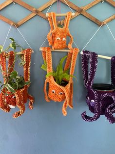 three orange and purple crocheted animals hanging from hooks on a wall next to a potted plant