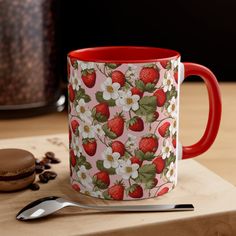 a red and white coffee mug sitting on top of a wooden table next to a cookie