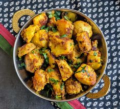 a metal bowl filled with cooked food on top of a colorful table cloth next to a spoon