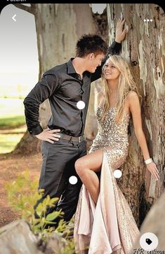 a man standing next to a woman in a dress near a tree and rock wall