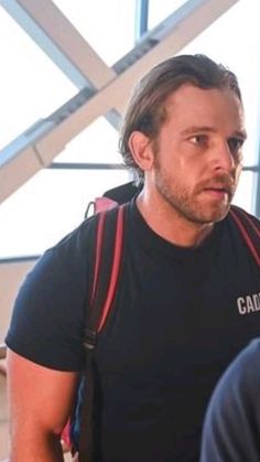 a man wearing a black shirt and carrying a red backpack in an airport with other people around him