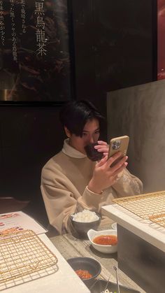 a man sitting at a table looking at his cell phone and eating rice with chopsticks in front of him