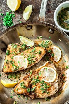 fish with lemons and herbs in a frying pan on a table next to silverware