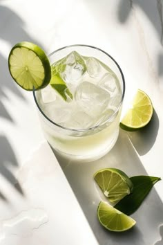 a glass filled with ice and limes on top of a white counter next to some cut up limes