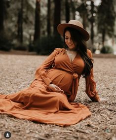 a pregnant woman sitting on the ground wearing a brown dress and hat with trees in the background