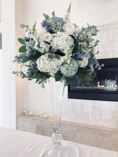 a vase filled with white and blue flowers sitting on top of a table next to a fireplace