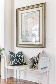 a white bench with two pillows on it in front of a framed painting and wooden floors