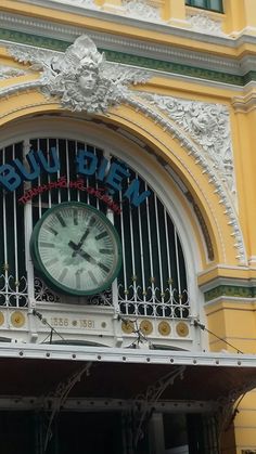 a large clock mounted to the side of a yellow and white building with ornate ironwork