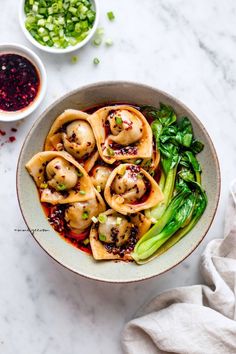 a bowl filled with dumplings and vegetables on top of a white table next to bowls of sauce