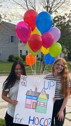 two girls holding up a sign that says are you up for hoco? with balloons