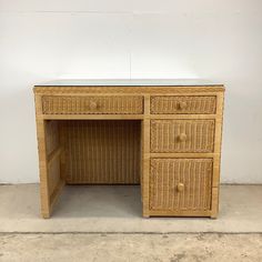 a wicker desk with two drawers and a glass top on concrete floor next to white wall