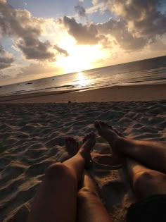 two people are laying on the beach watching the sun go down over the water and clouds