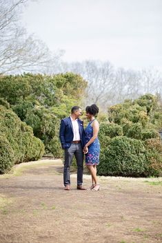 a man and woman standing next to each other on a dirt road in front of bushes