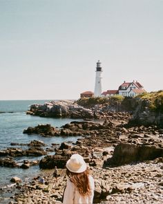 a woman standing on top of a rocky beach next to the ocean with a lighthouse in the background