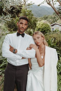 a man and woman standing next to each other in front of some bushes with trees