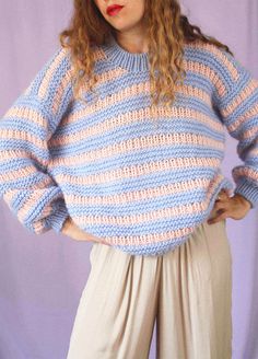 a woman wearing a blue and pink sweater with stripes on the sleeves, standing in front of a purple backdrop