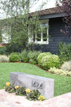 a cement sign sitting in the middle of a yard next to some bushes and flowers