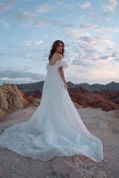 a woman standing on top of a rocky hill wearing a white wedding dress with ruffled sleeves