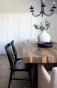 a dining room table with chairs and a vase on top of it, in front of a white paneled wall