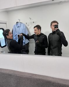 three men in wetsuits are looking at their cell phones while standing in front of a mirror