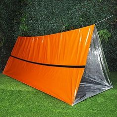 an orange tent sitting on top of green grass next to a bush with trees in the background