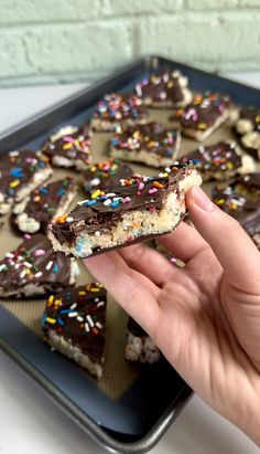 a person holding up a piece of chocolate and sprinkles cookie bar on a baking sheet