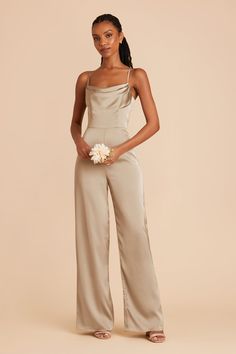 a woman standing in front of a beige background wearing a jumpsuit and holding a white flower