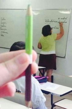 a hand holding a pencil in front of a whiteboard with writing on it and another person standing at the back