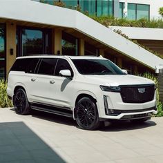 a white suv is parked in front of a large building with plants and bushes around it