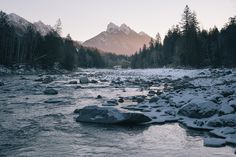 a river running through a forest filled with snow covered rocks and trees in the background