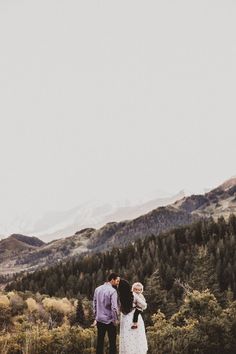 a man and woman standing on top of a grass covered field next to trees with mountains in the background