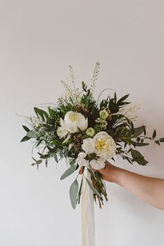 a person holding a bouquet with white flowers and greenery in their hand, against a wall