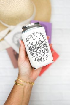 a person holding a can of beer in their hand with hats on the table behind it