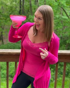 a woman holding a pink drink in her right hand and pointing to the side while standing on a deck