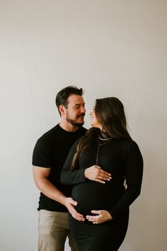 a pregnant couple cuddles in front of a white wall