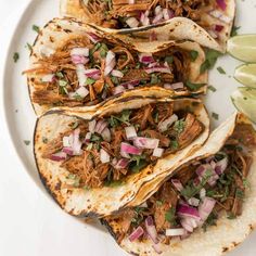 three shredded pork tacos with onions and cilantro on a white plate next to lime wedges