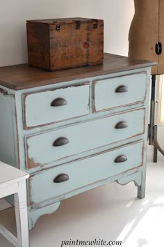 an old blue dresser with drawers and a wooden box on it's top, next to a white chair